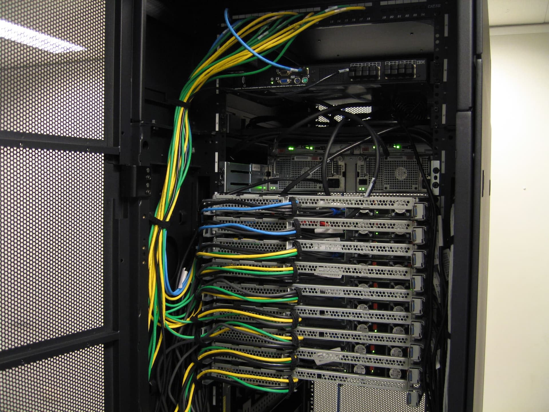 Armadio rack aperto con una pila di server e cavi di rete gialli, verdi e blu raccolti in ordine, in un locale tecnico luminoso.