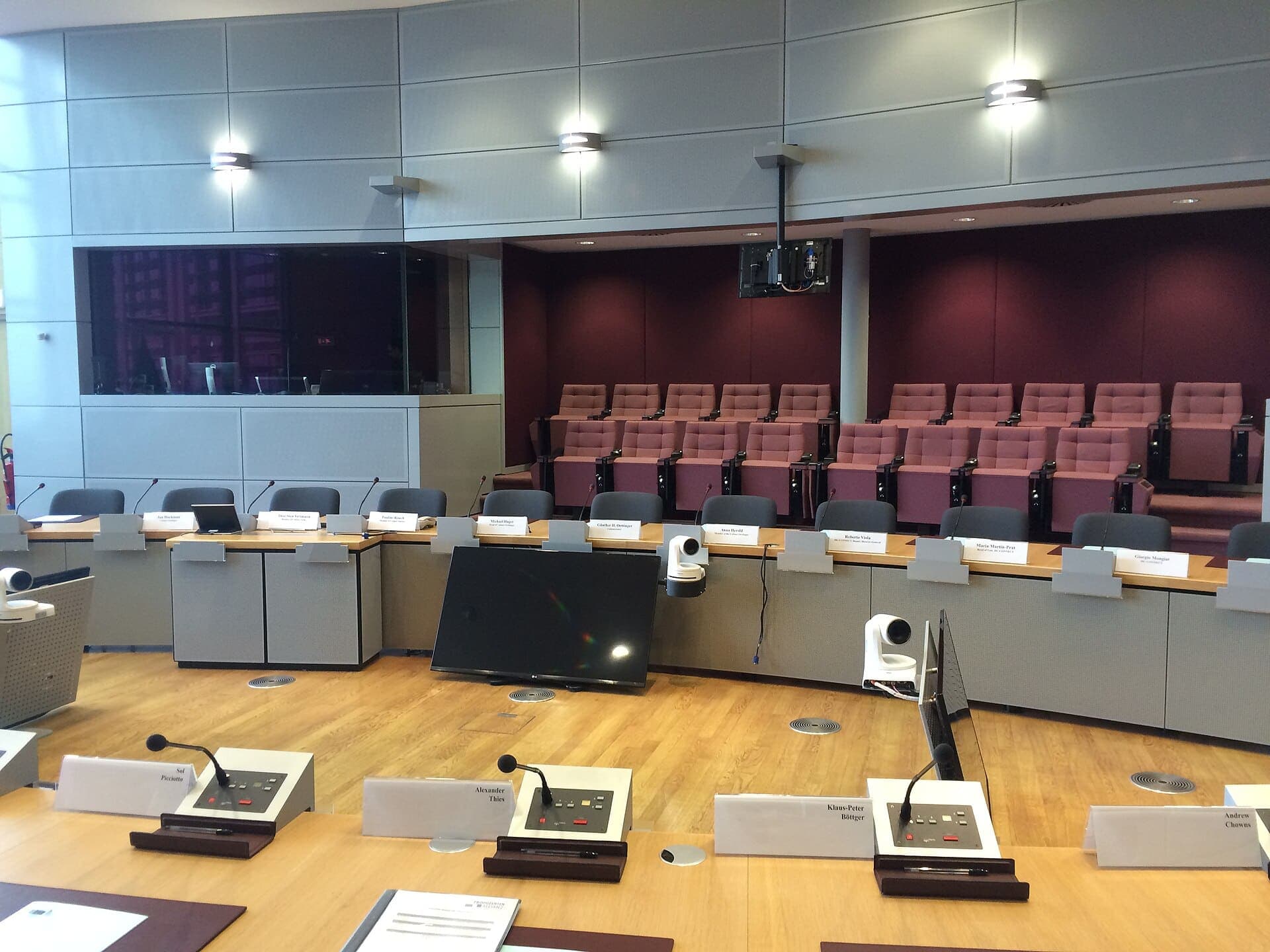 Sala riunioni con tavoli in legno chiaro e microfoni da conferenza davanti ai posti, cartellini dei partecipanti e telecamere sullo sfondo.