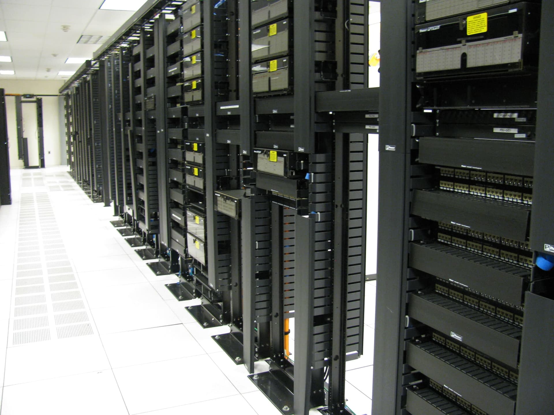 Corridoio di un data center con file di armadi rack neri su pavimento sopraelevato bianco, con cablaggi e pannelli di rete.